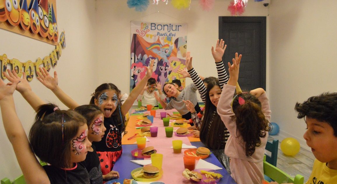 Children with painted faces raising their hands at a party table set with burgers