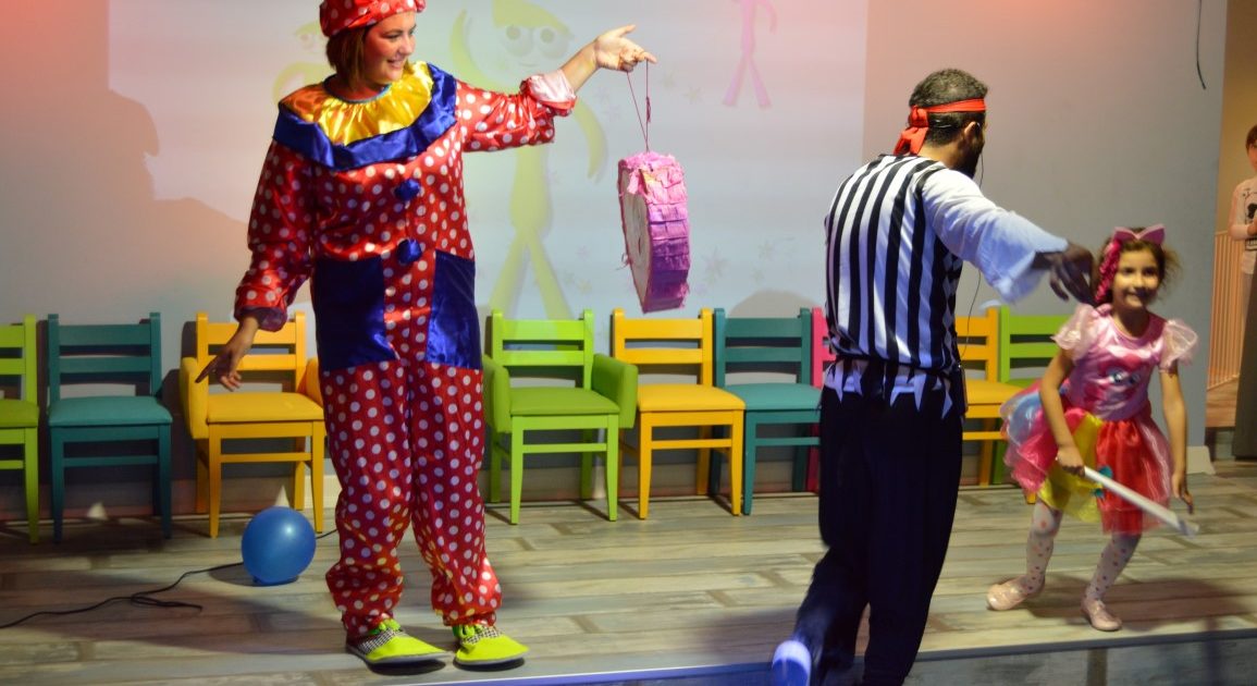 A clown holding a piñata on a string on stage, with an entertainer and a child waiting with a stick