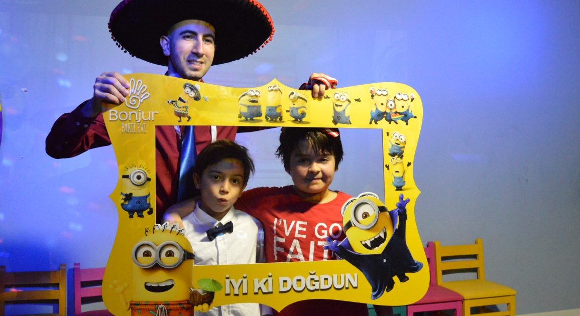 Two children posing with a yellow “Happy birthday” photo frame, with an entertainer in a wide-brimmed hat