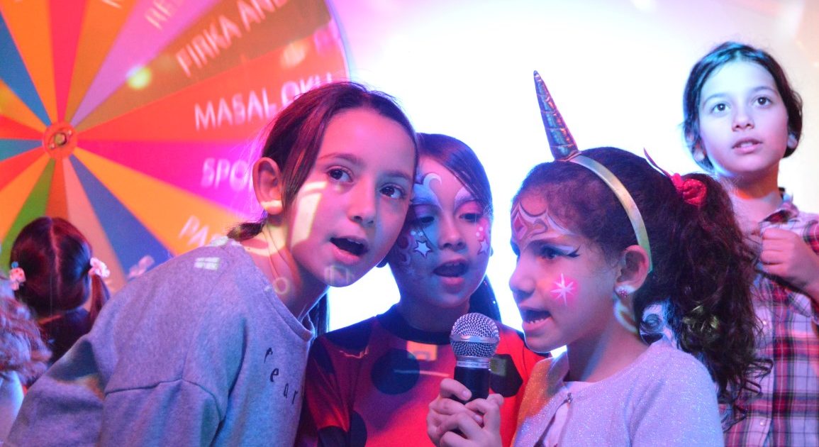 Children with painted faces singing into a microphone together under coloured lights