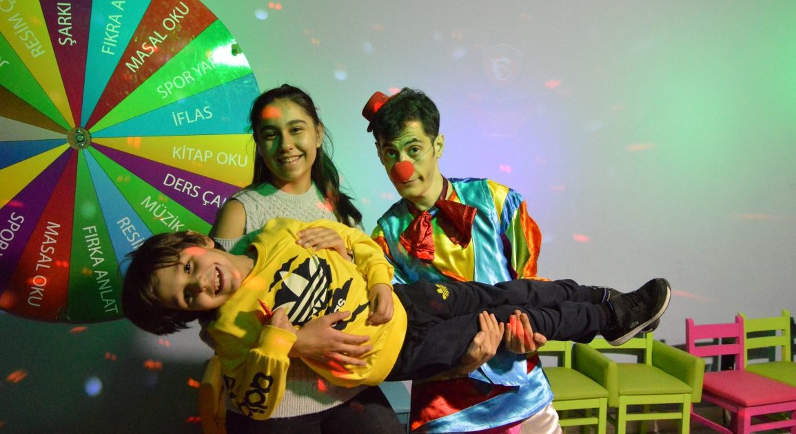 A clown and a girl carrying a laughing child in their arms under disco lights, in front of a colourful wheel