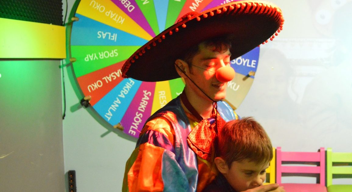 A clown in a wide-brimmed hat and a red nose with a child in front of the colourful game wheel