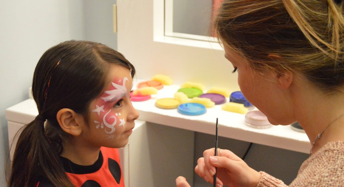 A child having star patterns painted on her face with a brush, with colourful face paints behind her