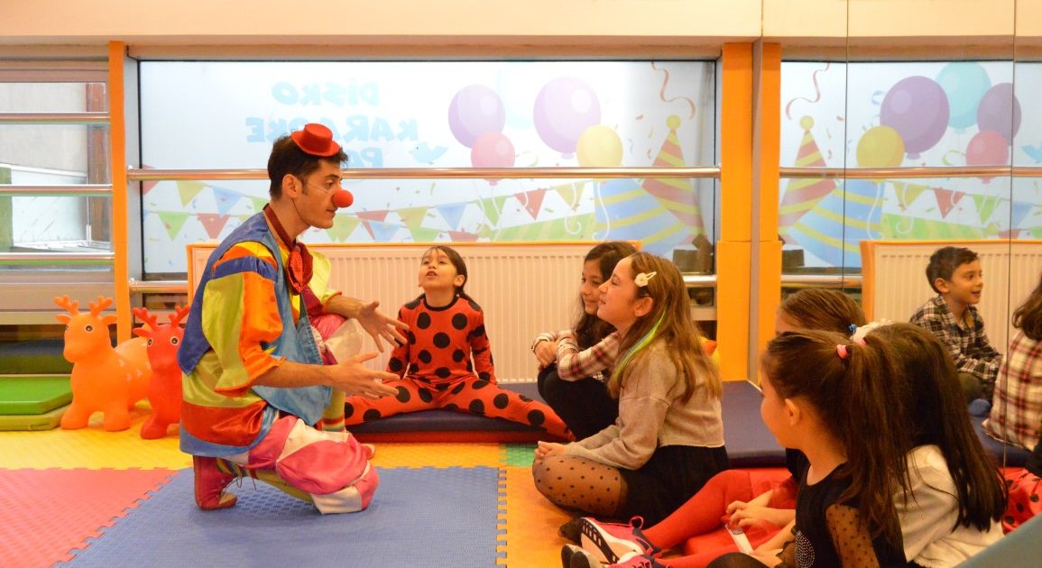 Children sitting on the floor of the play area around a clown, listening to him