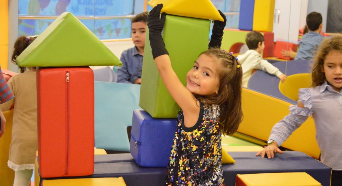 A smiling girl building a tower from soft blocks, with other children playing around her
